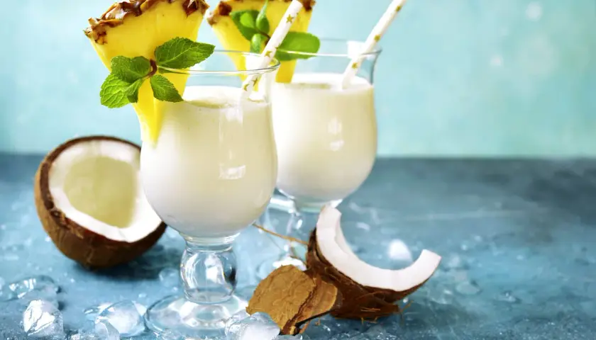 Zwei cremige Malibu Cocktails mit Ananas und Minze, umgeben von Kokosnuss und Eis.