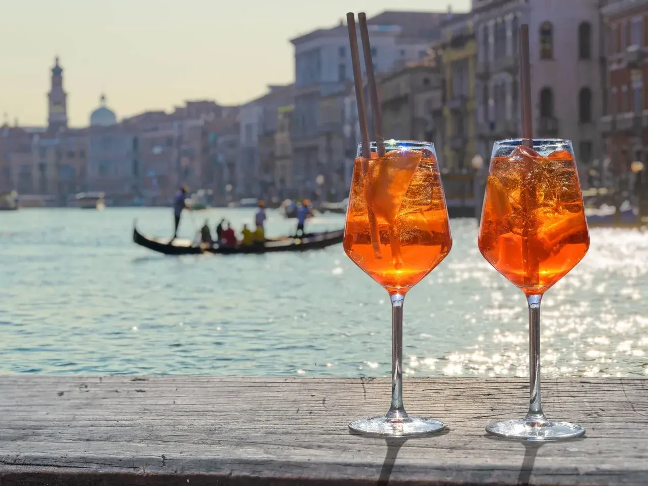 Zwei Aperol Spritz mit Eis und Orangenscheiben stehen auf einer Holzplanke. Im Hintergrund Venedig mit Gondel. Perfekt für Aperol Sekt trocken oder halbtrocken.
