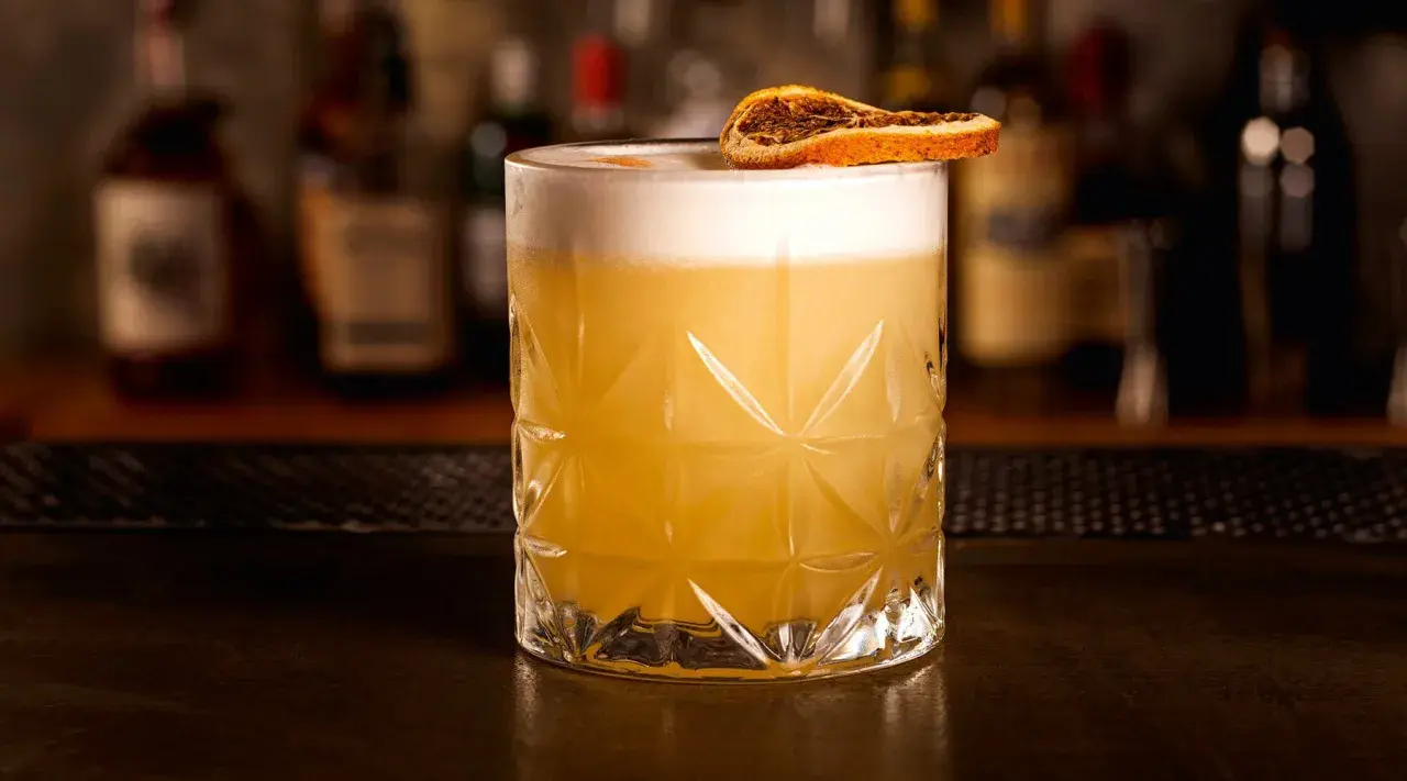 Ein erfrischender Grappa Cocktail mit cremigem Schaum und einer getrockneten Orangenscheibe, serviert in einem Glas mit geometrischem Muster.