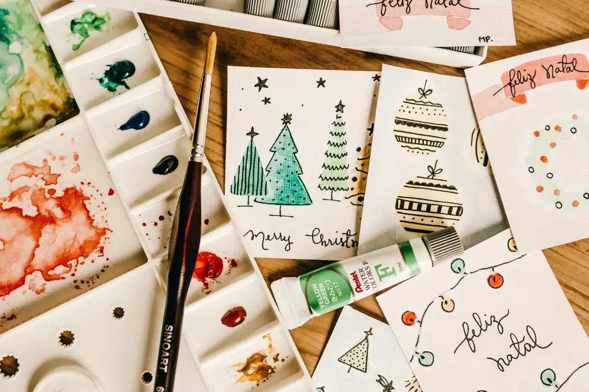 Bunte Aquarellfarben, Pinsel und selbstgemachte Weihnachtskarten mit Bäumen und Kugeln. Perfekt für eine DIY Weihnachtskarte!
