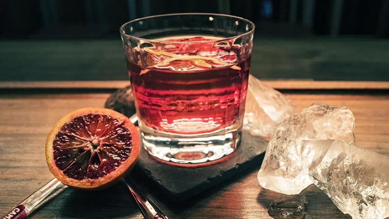 Ein Glas mit roten Cocktails, garniert mit Orangenzesten, neben einer Blutorange und Eiswürfeln auf einem Holztisch.