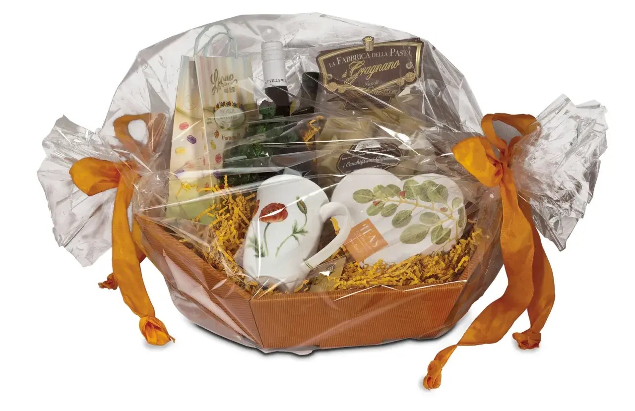 Ein liebevoll zusammengestelltes Geschenk, das mit Folie und orangefarbenen Bändern verpackt ist. Darin befinden sich Tassen, Pasta und weitere Leckereien.