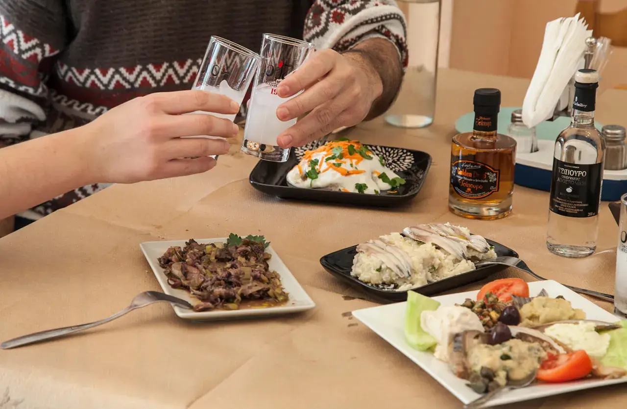 Zwei Gläser mit milchigem Ouzo-Getränk werden angestoßen. Dazu gibt es verschiedene griechische Mezedes und kleine Flaschen Ouzo.