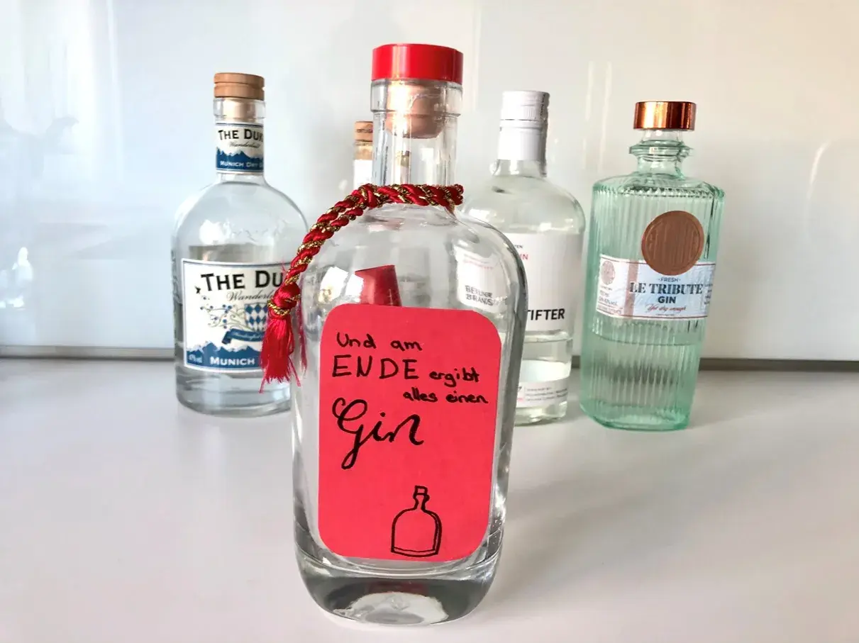 Ein roter Zettel mit der Aufschrift "Und am Ende ergibt alles einen Gin" ist an einer leeren Flasche befestigt. Mehrere Gin-Flaschen stehen im Hintergrund.