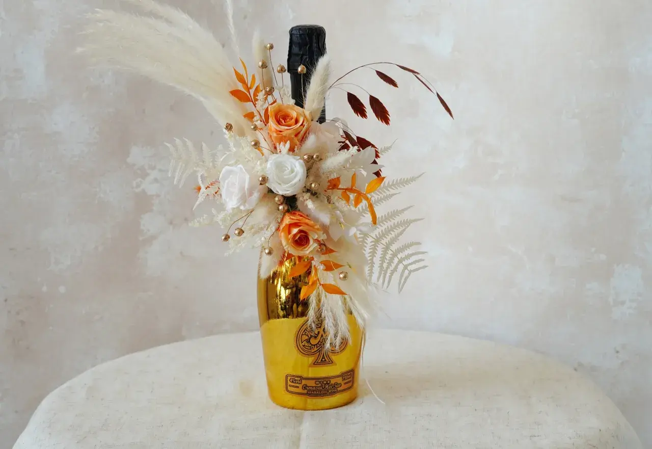 Goldene Weinflasche mit Blumen verpackt. Ein Strauß aus getrockneten Blumen, Rosen und Pampasgras schmückt die Flasche.