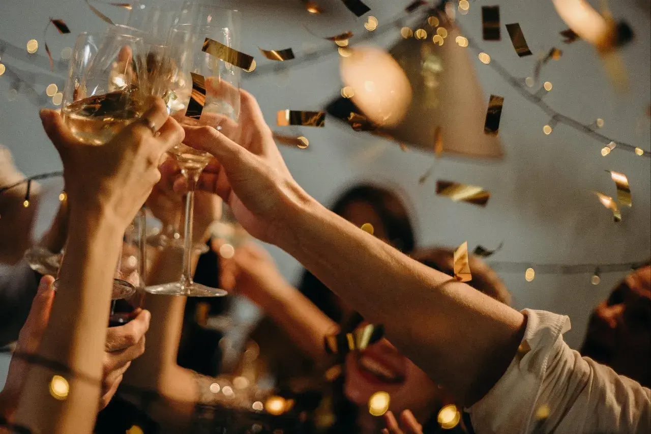 Hände stoßen mit Gläsern an, Konfetti fällt. Ein fröhlicher Anlass, vielleicht ein Jubiläum, wird gefeiert.