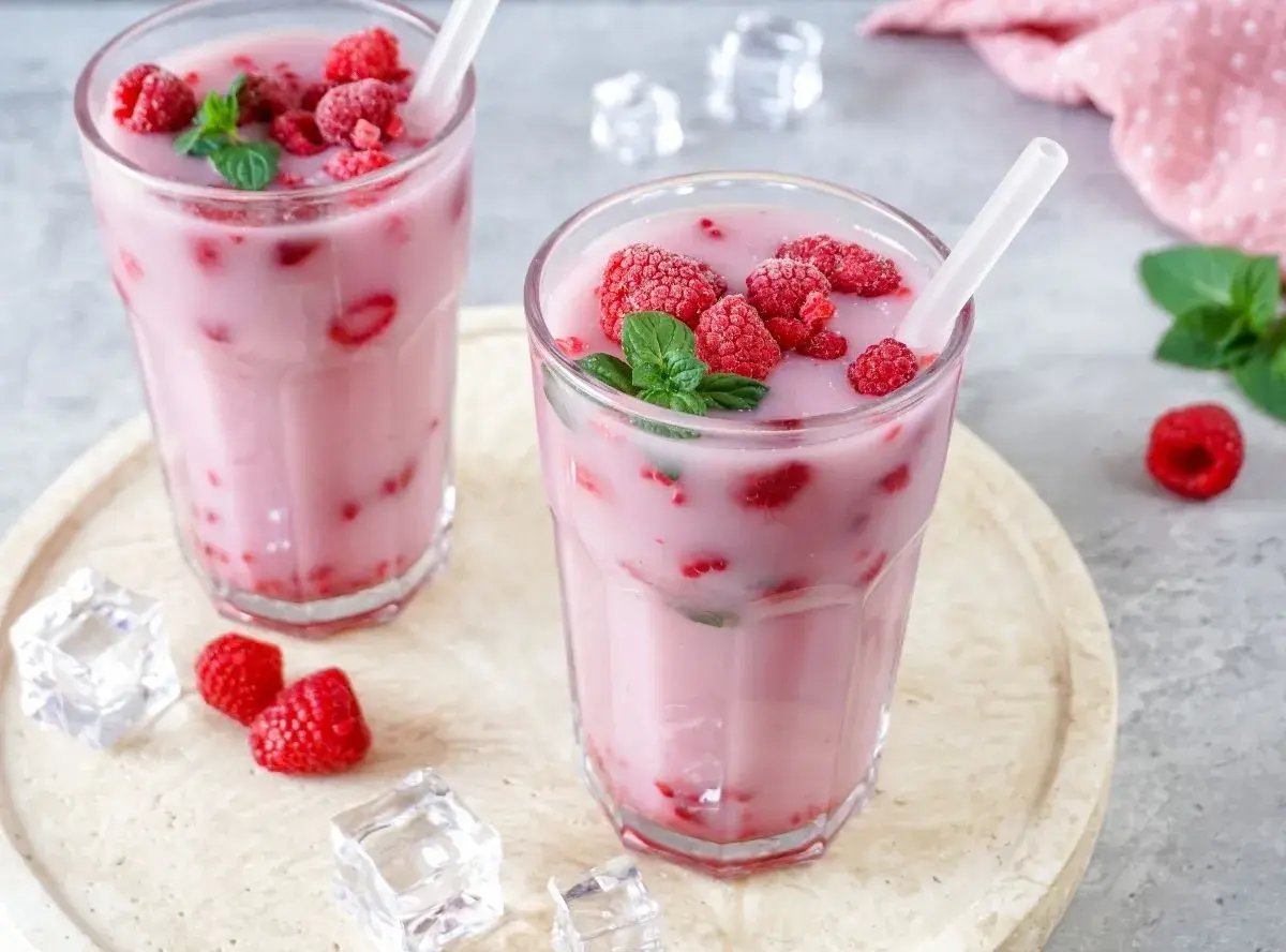 Zwei Gläser mit einem erfrischenden, pinken Drink, gefüllt mit Himbeeren und garniert mit Minze. Eiswürfel und frische Himbeeren liegen daneben.