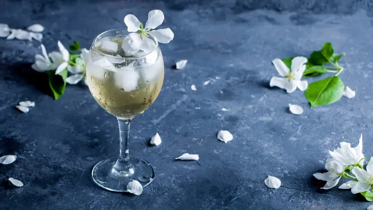 Ein Glas mit einem erfrischenden, eisgekühlten Getränk, garniert mit einer weißen Blüte. Zarte Blütenblätter liegen auf dem dunklen Untergrund. Ein sommerlicher porto tonico.