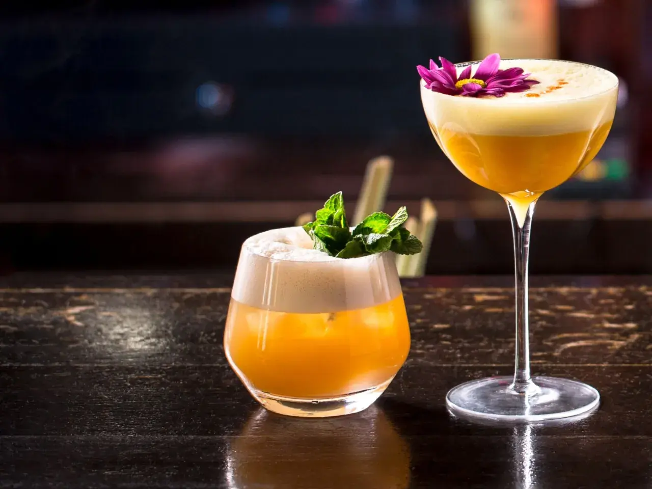 Zwei exquisite Brandy Cocktails, einer mit Minze, der andere mit einer lila Blüte garniert, stehen auf einem dunklen Holztisch.