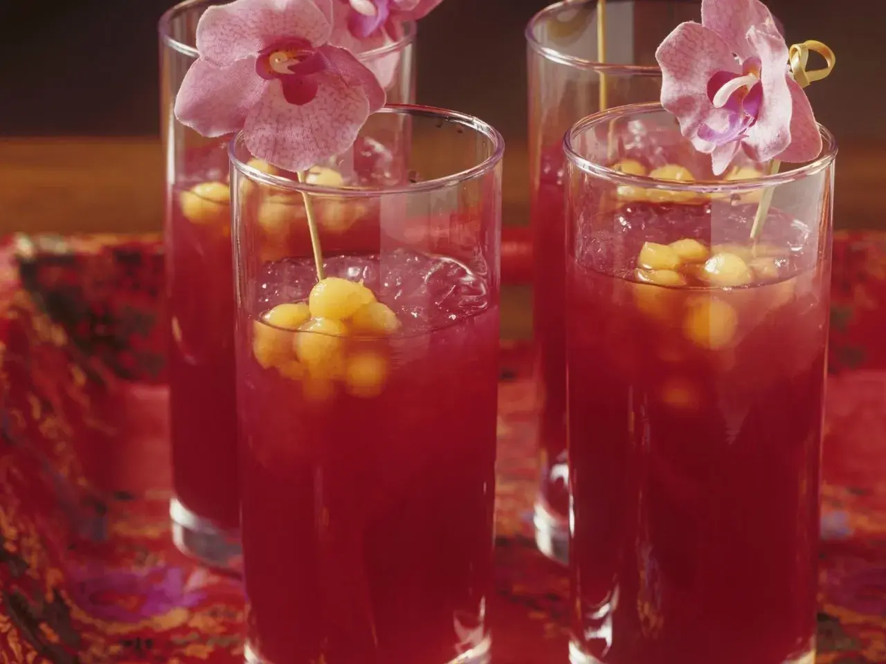 Erfrischende asiatische Cocktails mit Melonenbällchen und Orchideen auf einem roten, gemusterten Tuch.