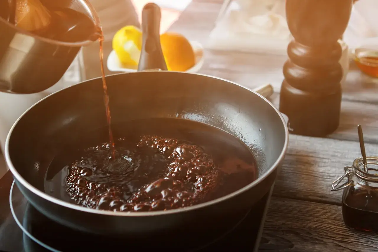 Dunkler Portwein zum Kochen wird in eine Pfanne gegossen, wo er zu köcheln beginnt. Zitrusfrüchte und ein Gewürzstreuer sind im Hintergrund zu sehen.