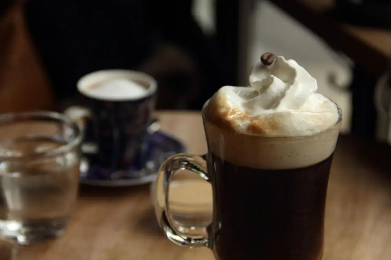 Ein Glas mit Kaffee und Sahne, garniert mit einer Kaffeebohne. Daneben ein kleiner Becher und ein Glas Wasser. Perfekt für einen Kaffee mit Schnaps.
