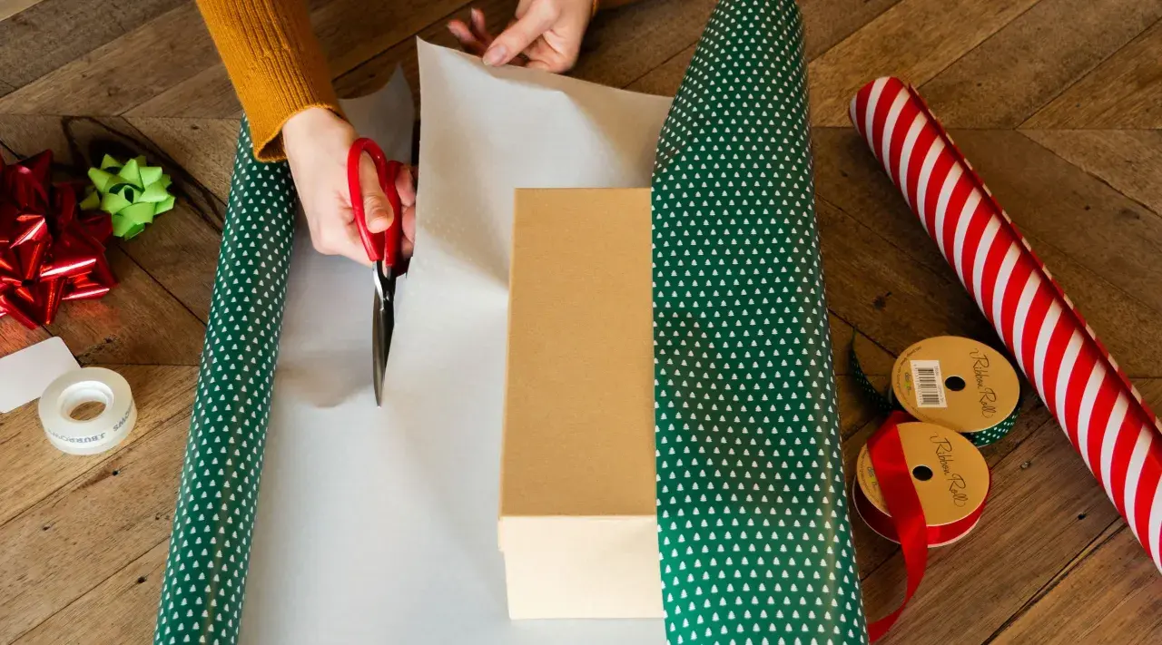Hände schneiden Geschenkpapier zu, um ein Geburtstagsgeschenk zu verpacken. Rote Schere, Geschenkband und Schleifen liegen bereit.