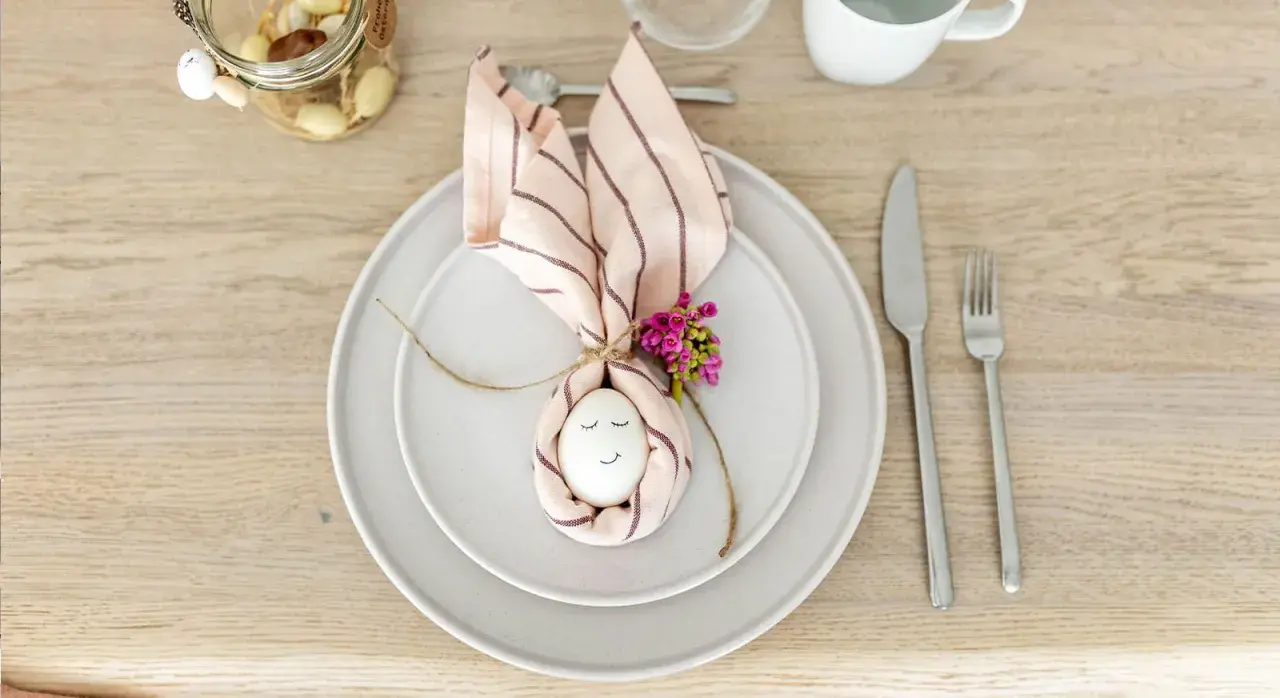 Niedliche Tischdeko Ostern einfach: Ein Ei mit Gesicht, umwickelt von einer Serviette als Hasenohren, mit kleinen Blüten verziert.