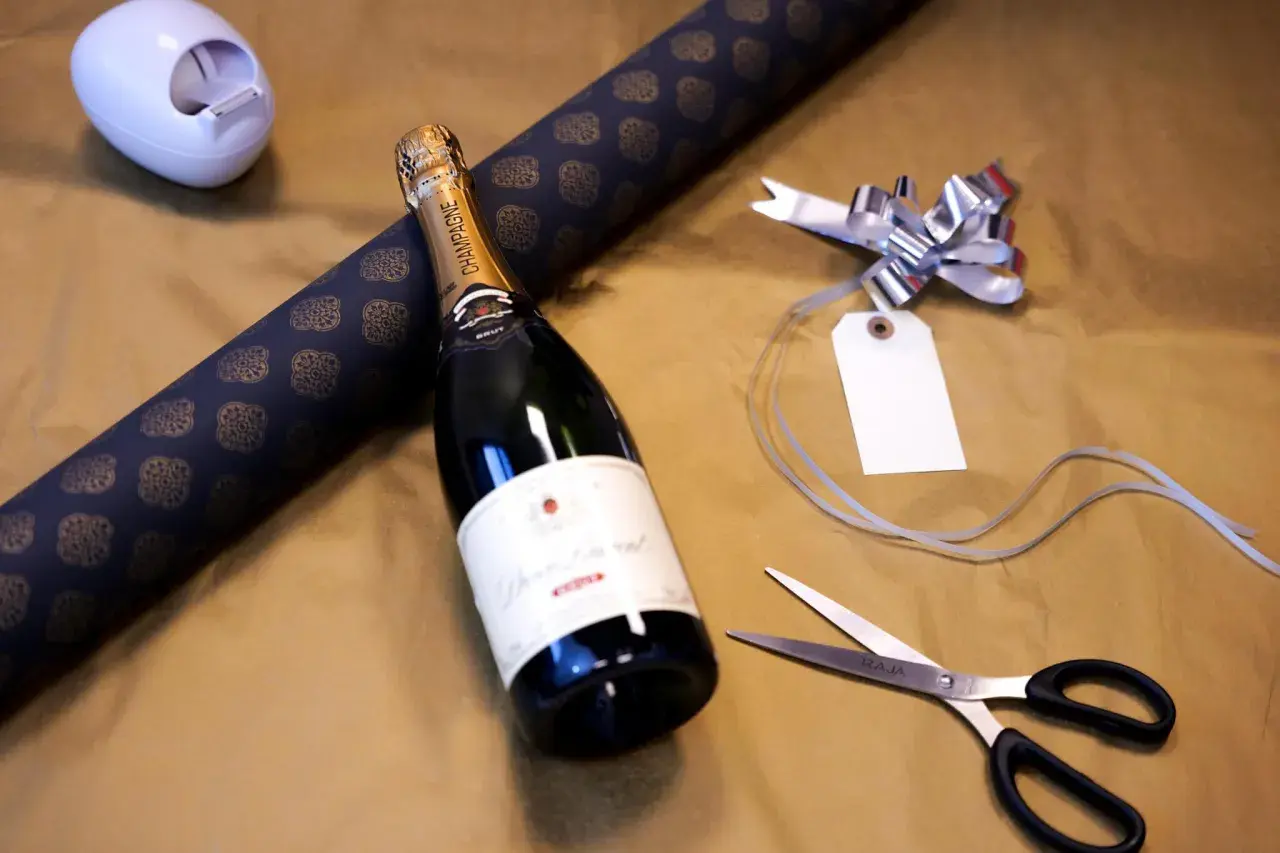 Vorbereitung, um mehrere Flaschen als Geschenk zu verpacken: Champagnerflasche, Geschenkpapier, Schere und Schleife liegen bereit.