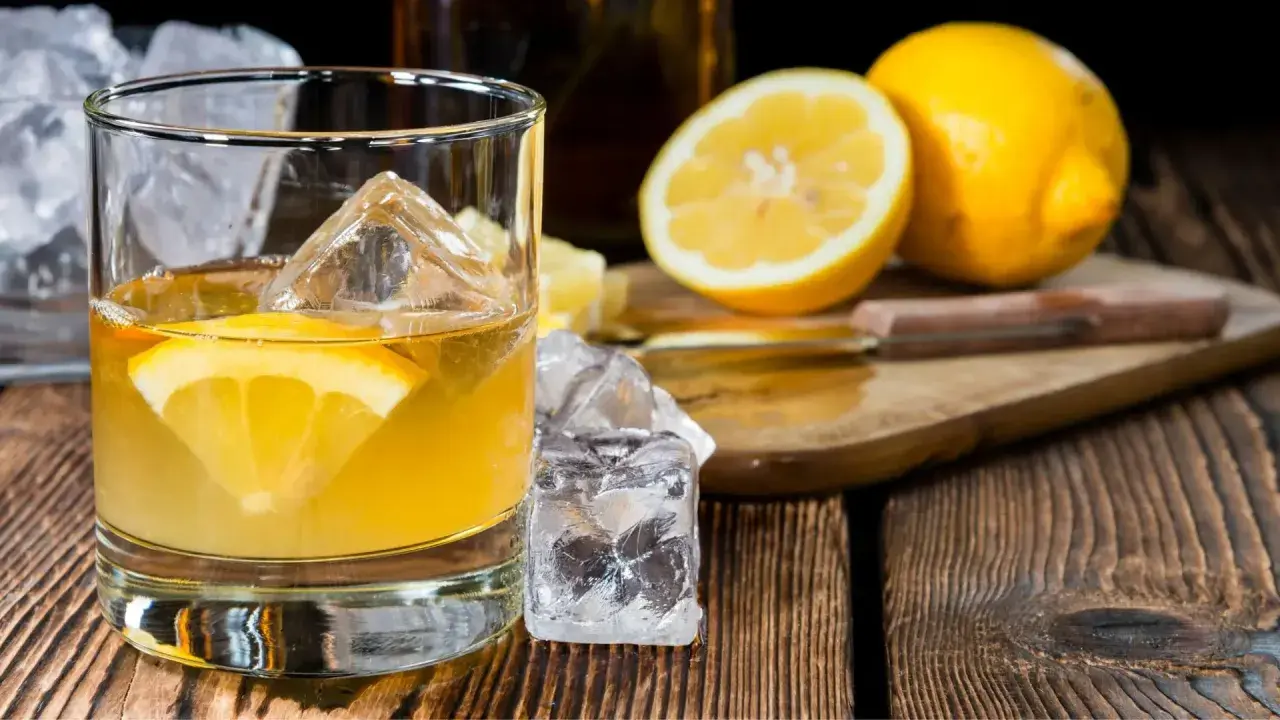 Ein erfrischender Penicillin Cocktail mit Eiswürfeln und Zitronenscheibe auf Holztisch. Daneben liegen frische Zitronen.