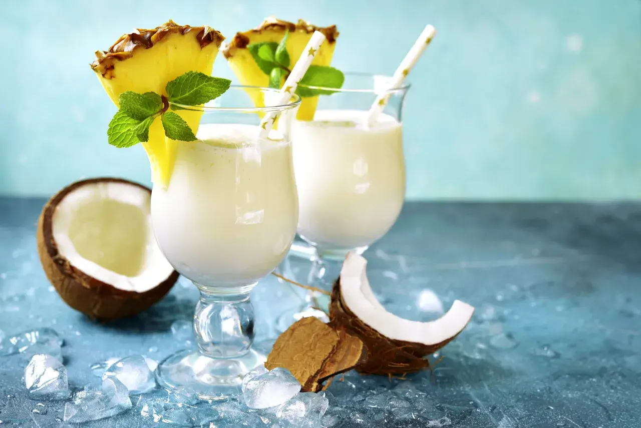 Zwei cremige Malibu Cocktails mit Ananas und Minze, umgeben von Kokosnuss und Eis.