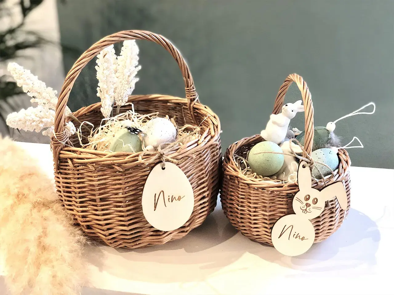 Zwei geflochtene Körbe mit Osterdeko: Eier, Federn, weiße Blumen und Holzanhänger mit Namen. Tolle Osterkorb Ideen für Kinder.