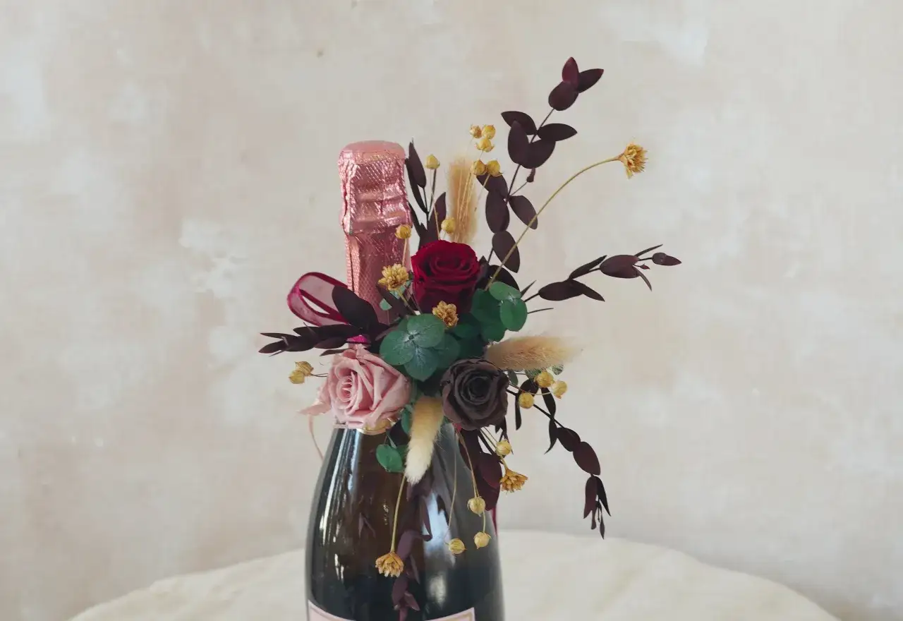 Eine Weinflasche mit Blumen verpacken: Ein Strauß aus Rosen, Pampasgras und dunklen Blättern schmückt den Hals einer Champagnerflasche.