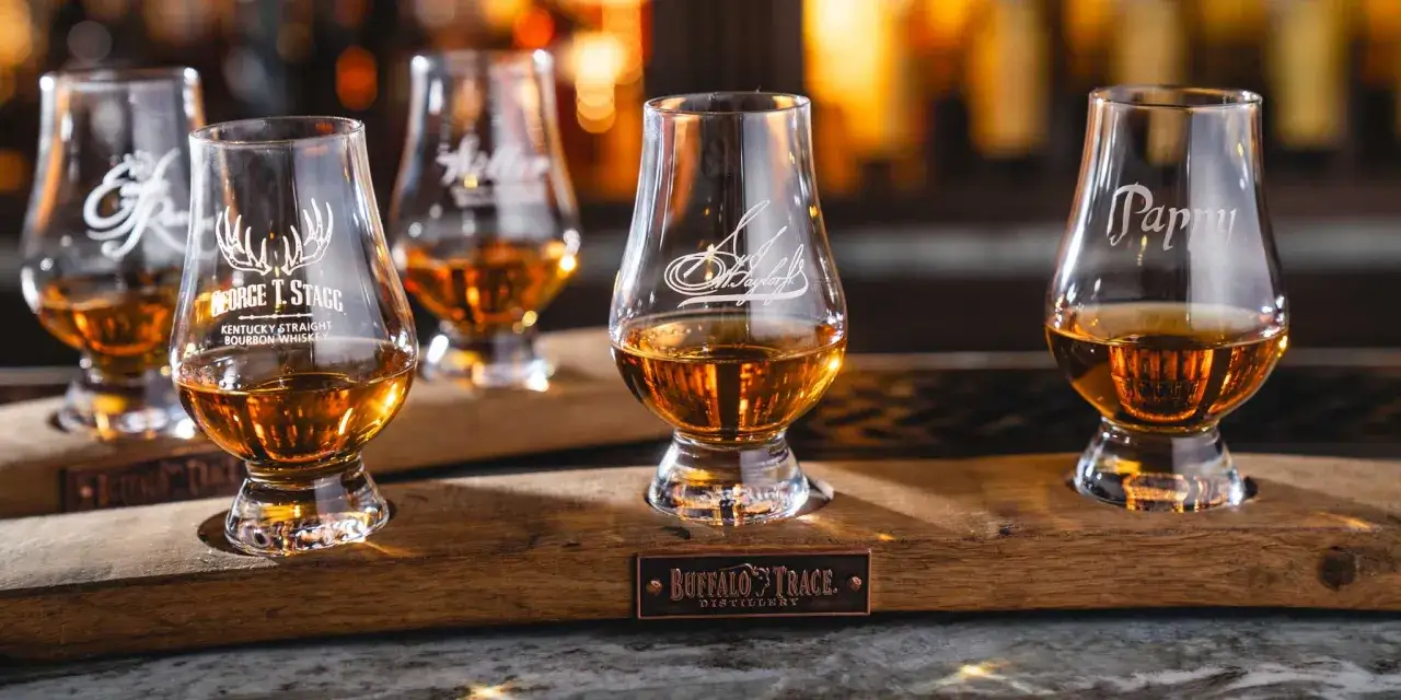 Vier Gläser mit bernsteinfarbenem Bourbon stehen auf einem Holzbrett. Jedes Glas trägt den Namen eines bekannten Bourbons, was die Vielfalt von was ist Bourbon zeigt.