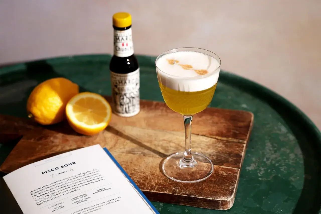 Ein Pisco Sour, ein klassischer Cocktail aus Chile, mit Zitronen, einem Fläschchen Angostura Bitter und einem Rezeptbuch.