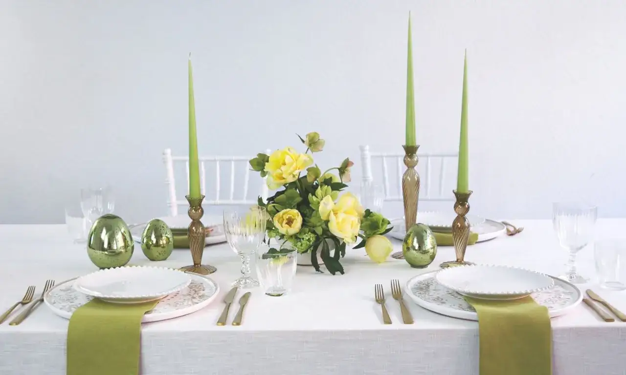 Einfache Tischdeko Ostern: Gr&uuml;ne Kerzen, goldene Eier und gelbe Blumen auf wei&szlig; gedecktem Tisch.