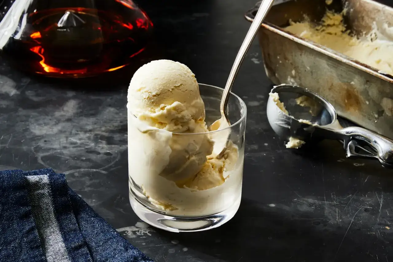 Cremiges Vanilleeis mit einem Hauch von Alkohol, serviert in einem Glas. Daneben ein Eisportionierer und eine Karaffe mit bernsteinfarbener Flüssigkeit.