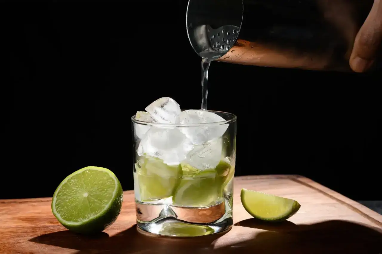 Eisw&uuml;rfel und Limettenspalten in einem Glas, w&auml;hrend eine Fl&uuml;ssigkeit aus einem Shaker gegossen wird. Das ist die Zubereitung eines Caipirinha, der mit Cachaca gemacht wird.
