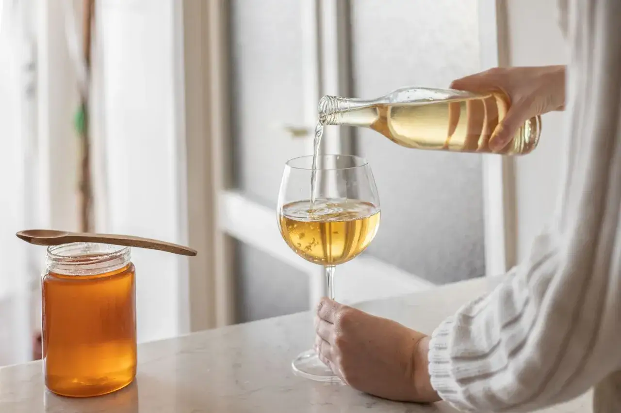 Ein Glas Met wird aus einer Flasche in ein Weinglas gegossen. Daneben steht ein Glas Honig mit einem Holzlöffel. So trinkt man Met!
