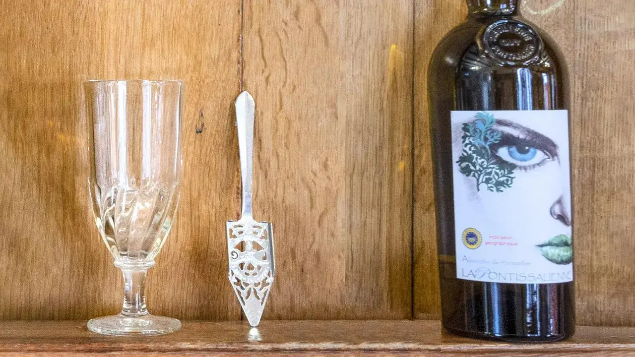 Ein Glas, ein L&ouml;ffel und eine Flasche Absinth. Die Flasche zeigt ein Auge und Lippen. Die Absinth Wirkung wird oft als berauschend beschrieben.