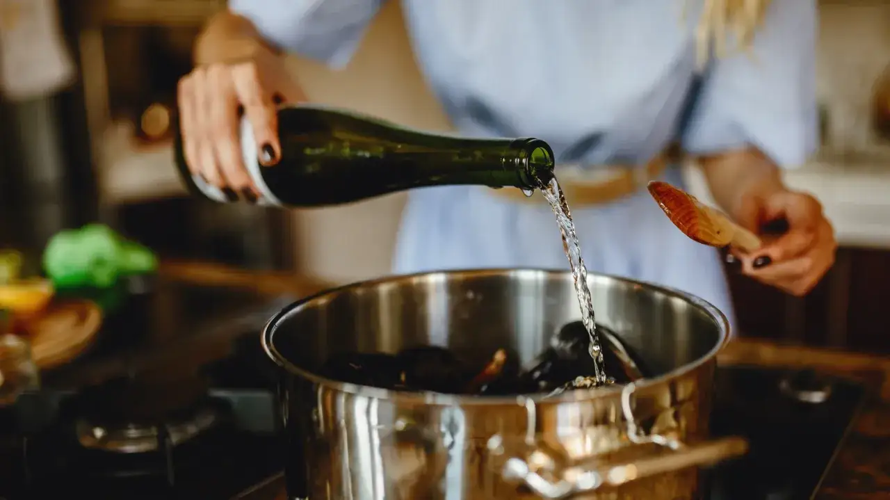 Frau gießt Weißwein als alkohol zum kochen in einen Topf mit Muscheln.