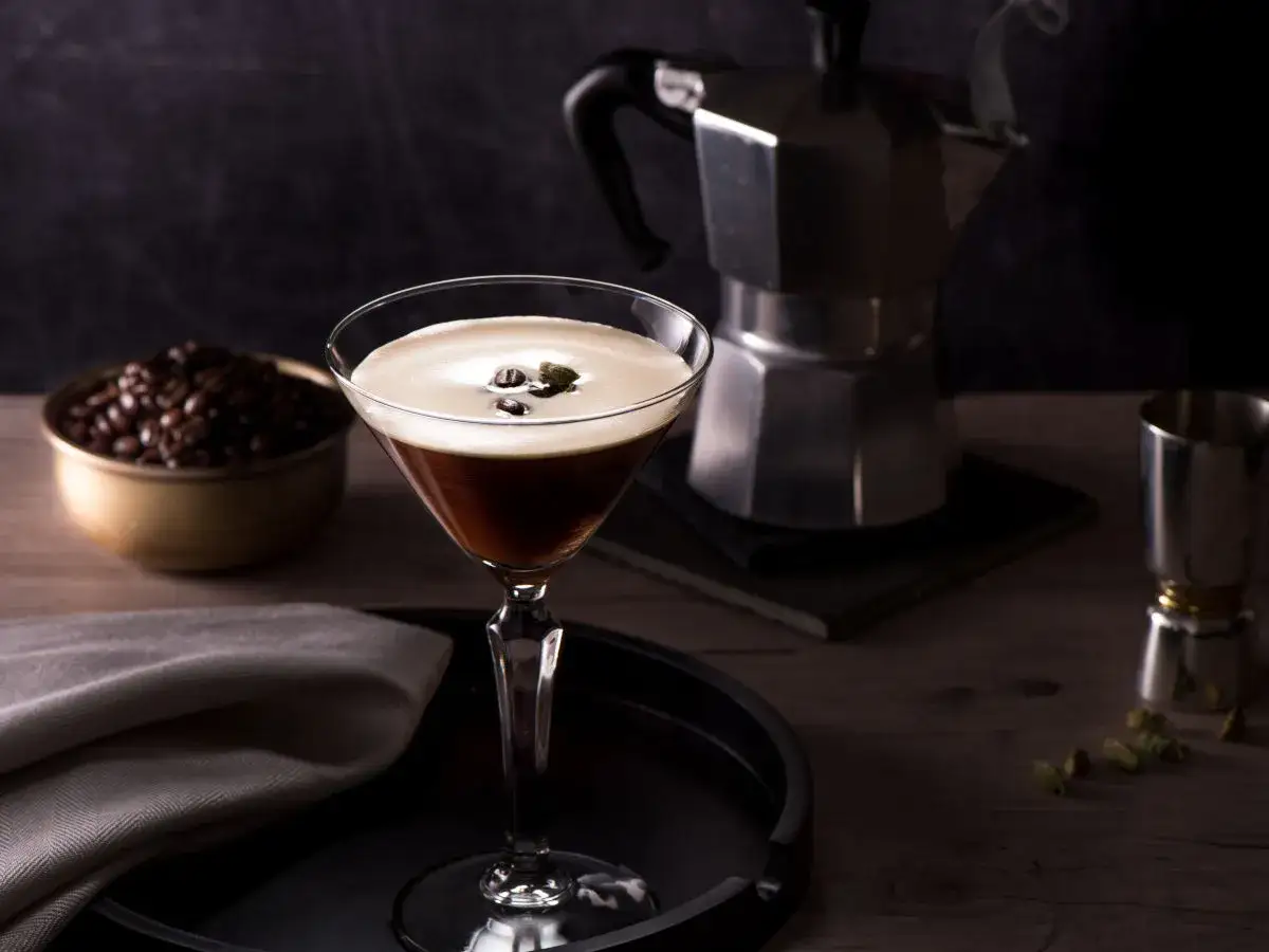 Ein eleganter Martini-Cocktail mit cremigem Schaum und Kaffeebohnen. Daneben eine Moka-Kanne, Kaffeebohnen und ein Schnapsglas. Perfekt für einen Kaffee mit Schnaps.