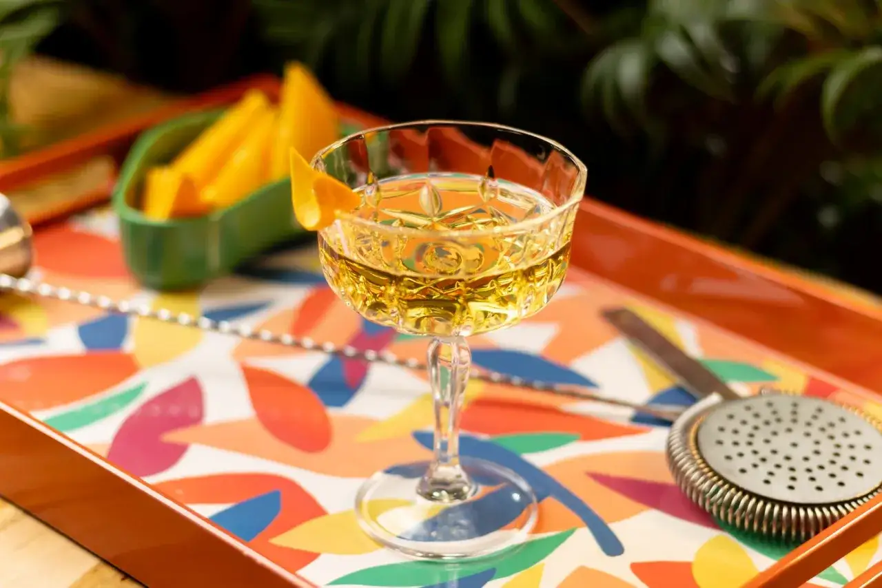 Ein goldener Cocktail mit Orangenzeste, bereit f&uuml;r herbe Cocktails. Daneben liegen Orangenst&uuml;cke und Bar-Utensilien auf einem bunten Tablett.