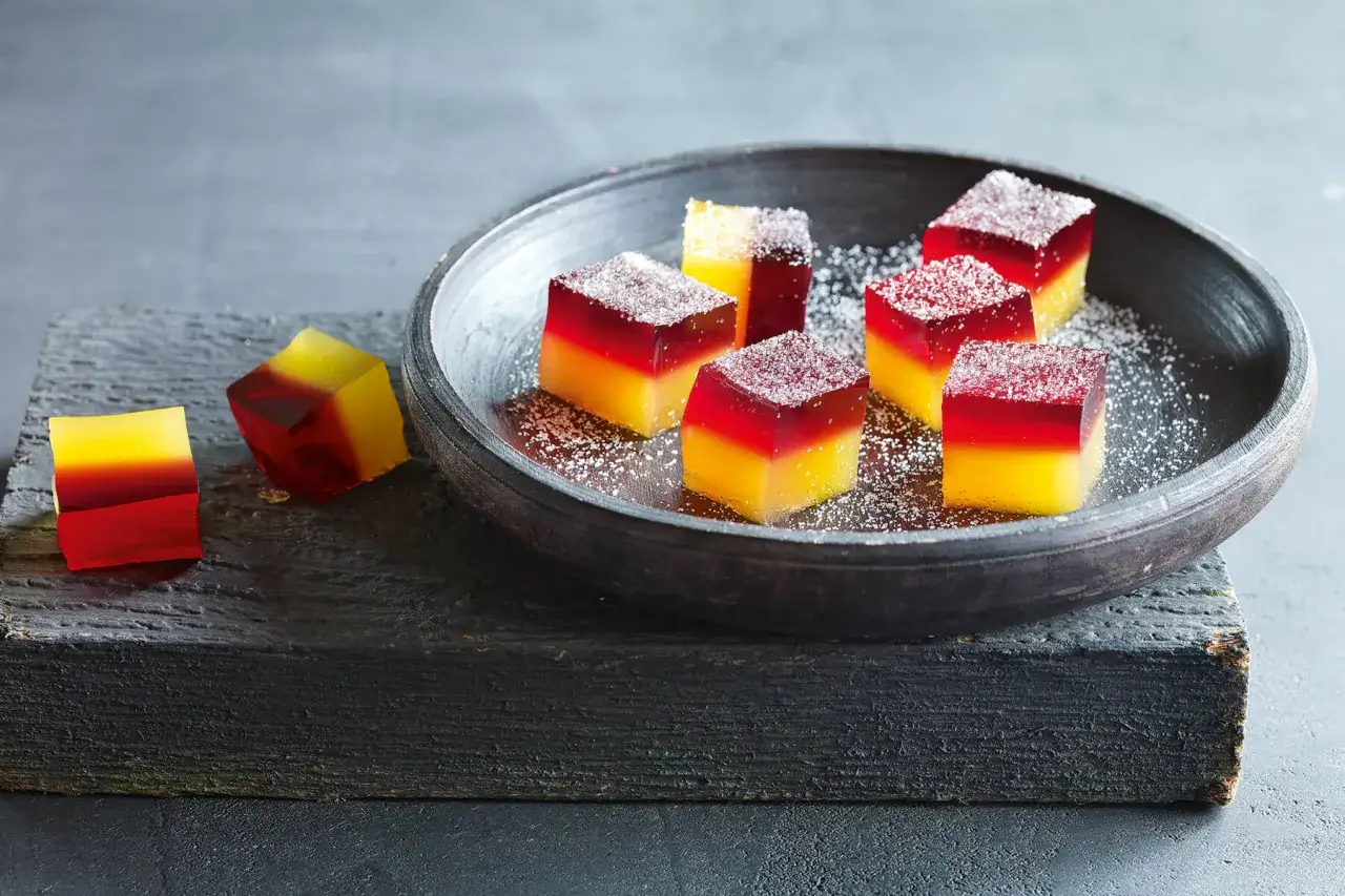 Bunte, geschichtete Jelly Shots, bestäubt mit Puderzucker, liegen in einer Schale und auf einem Holzbrett.