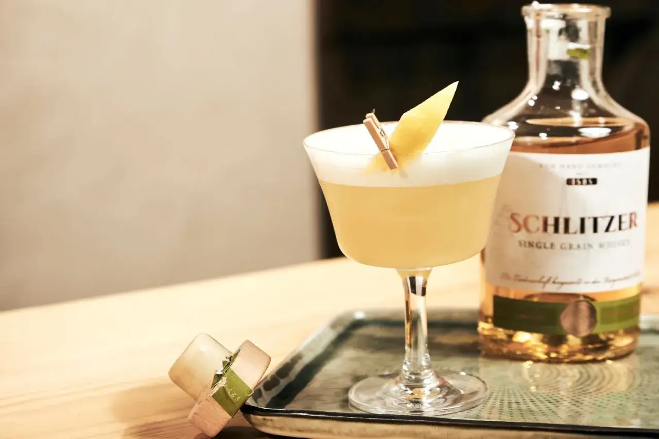 Ein erfrischender Whiskey Sour mit Zitronenzeste, daneben eine Flasche Schlitzer Single Grain Whisky.