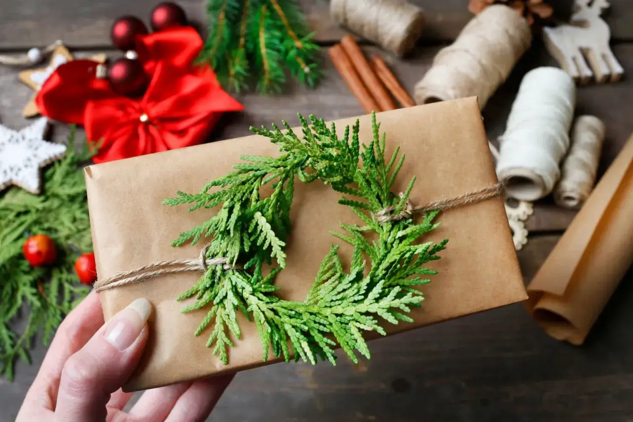 Geschenke verpacken Weihnachten Ideen: Ein braunes Geschenk mit Tannenzweig-Kranz, umgeben von Weihnachtsschmuck und Garnrollen.