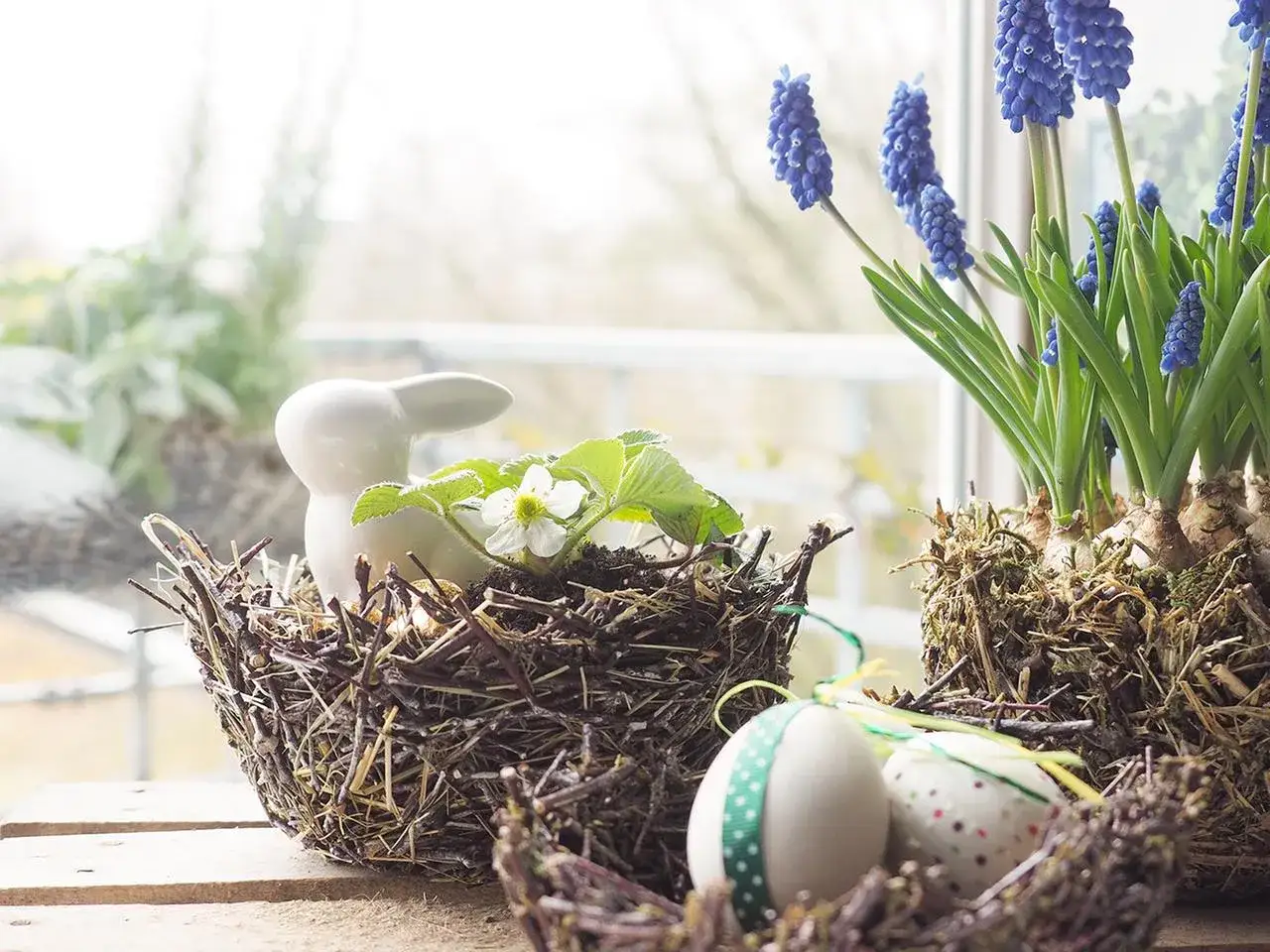 Osterkorb Ideen: Ein Keramik-Hase sitzt in einem Nest aus Zweigen, daneben blaue Traubenhyazinthen und verzierte Ostereier.