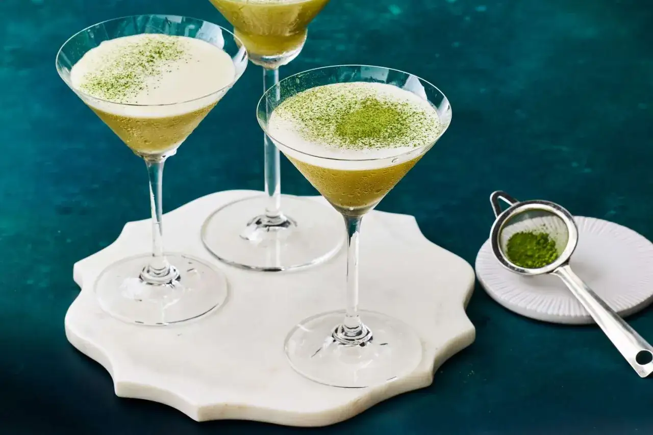Drei elegante Gläser mit asiatischen Cocktails, bestäubt mit grünem Matcha-Pulver. Ein kleines Sieb mit Matcha liegt daneben.