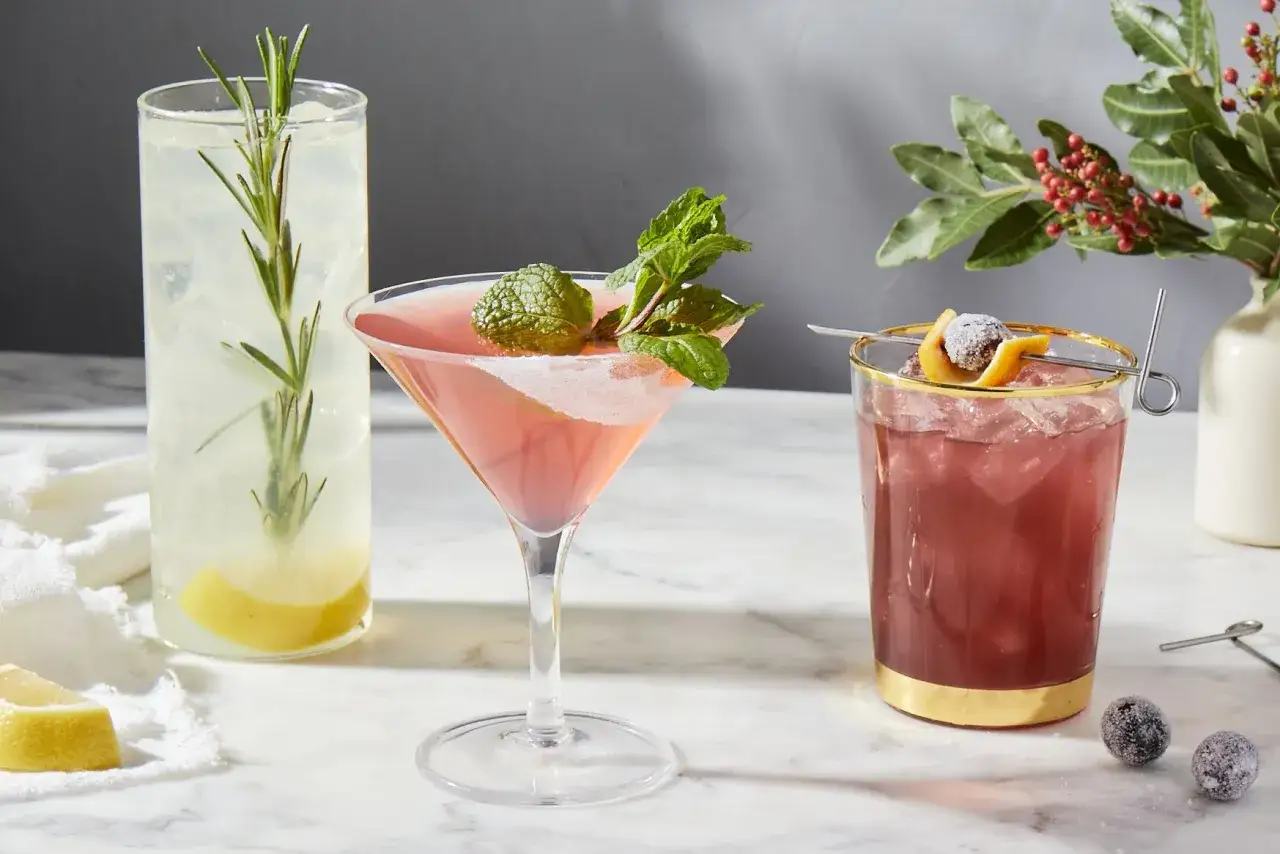 Drei bunte Cocktails mit Garnituren aus Minze, Rosmarin und Fr&uuml;chten stehen bereit f&uuml;r einen sommerlichen Genuss.
